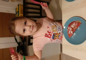 Dziewczynka trzymająca nóż i widelec w rękach, przed nią talerz z czerwonym naleśniczkiem
